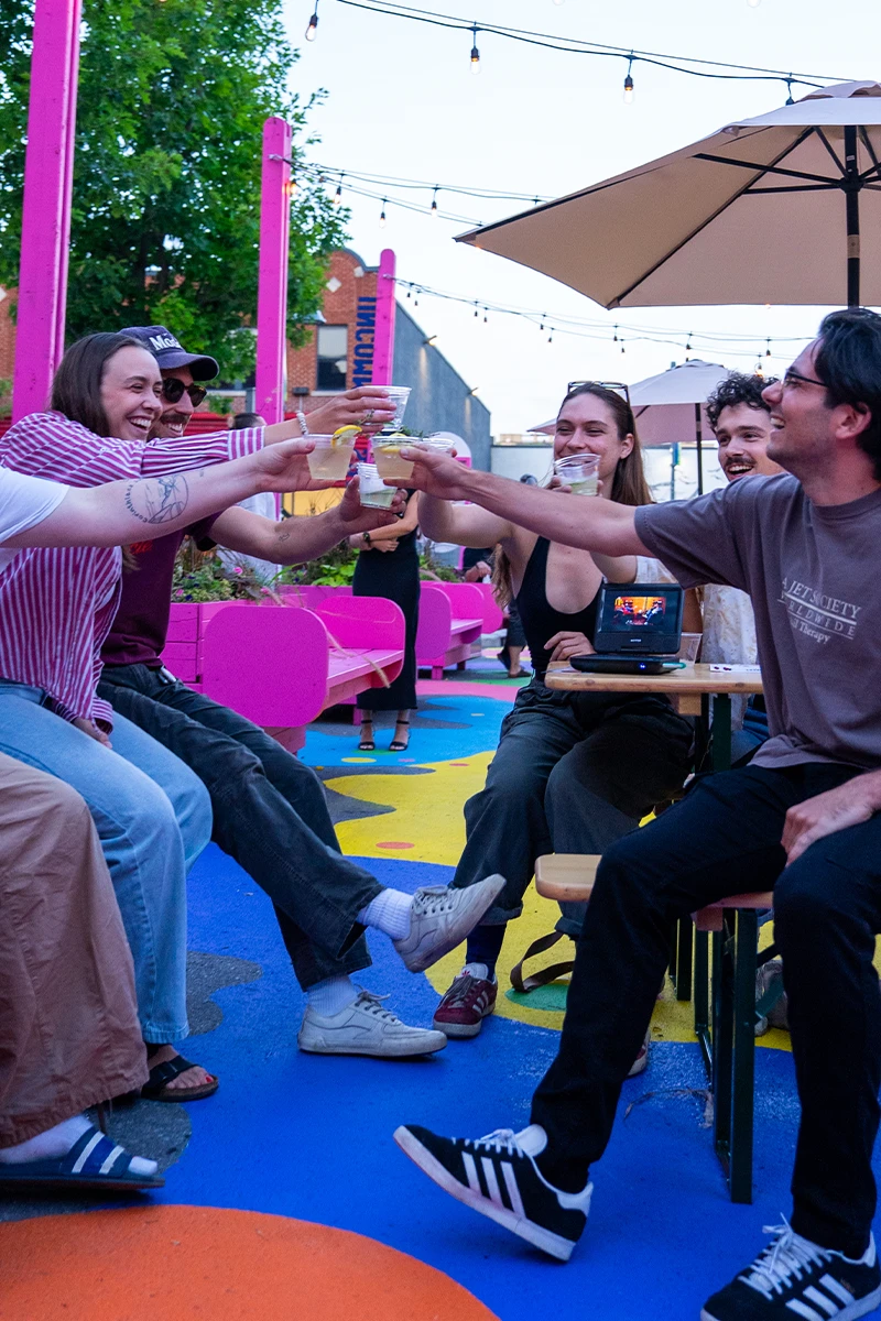 Six friends leaning in to clink drinks at an outdoor patio with pink benches and string lights overhead.
